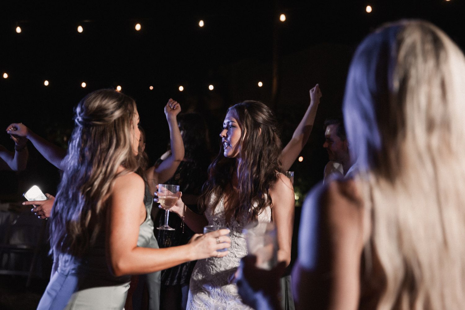 Bridesmaids and guests dance joyfully under string lights at lively outdoor wedding reception at night.