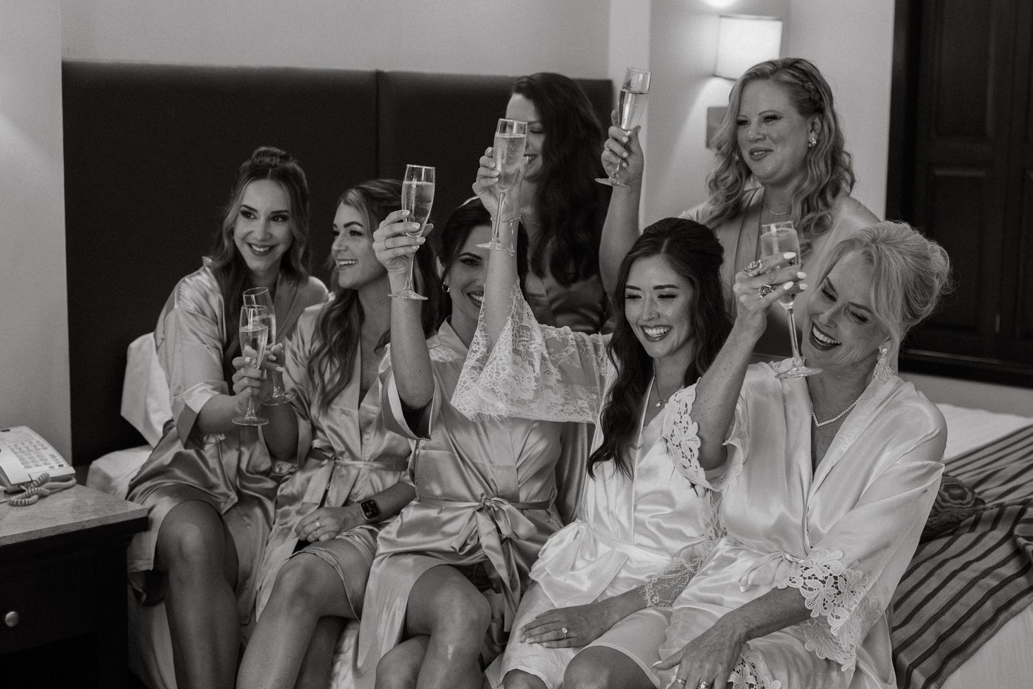 Bridesmaids in satin robes toasting with champagne glasses during wedding morning preparations in black and white photo.