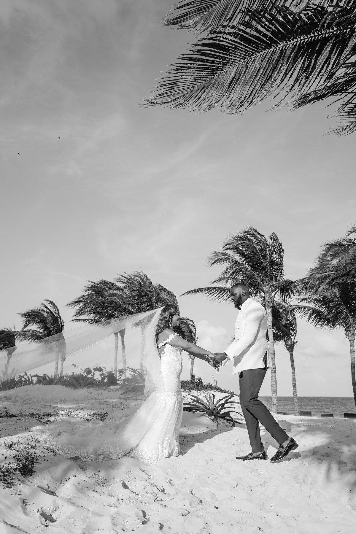 Black and white photo of bride and groom dancing on a sandy beach with palm trees in the wind.