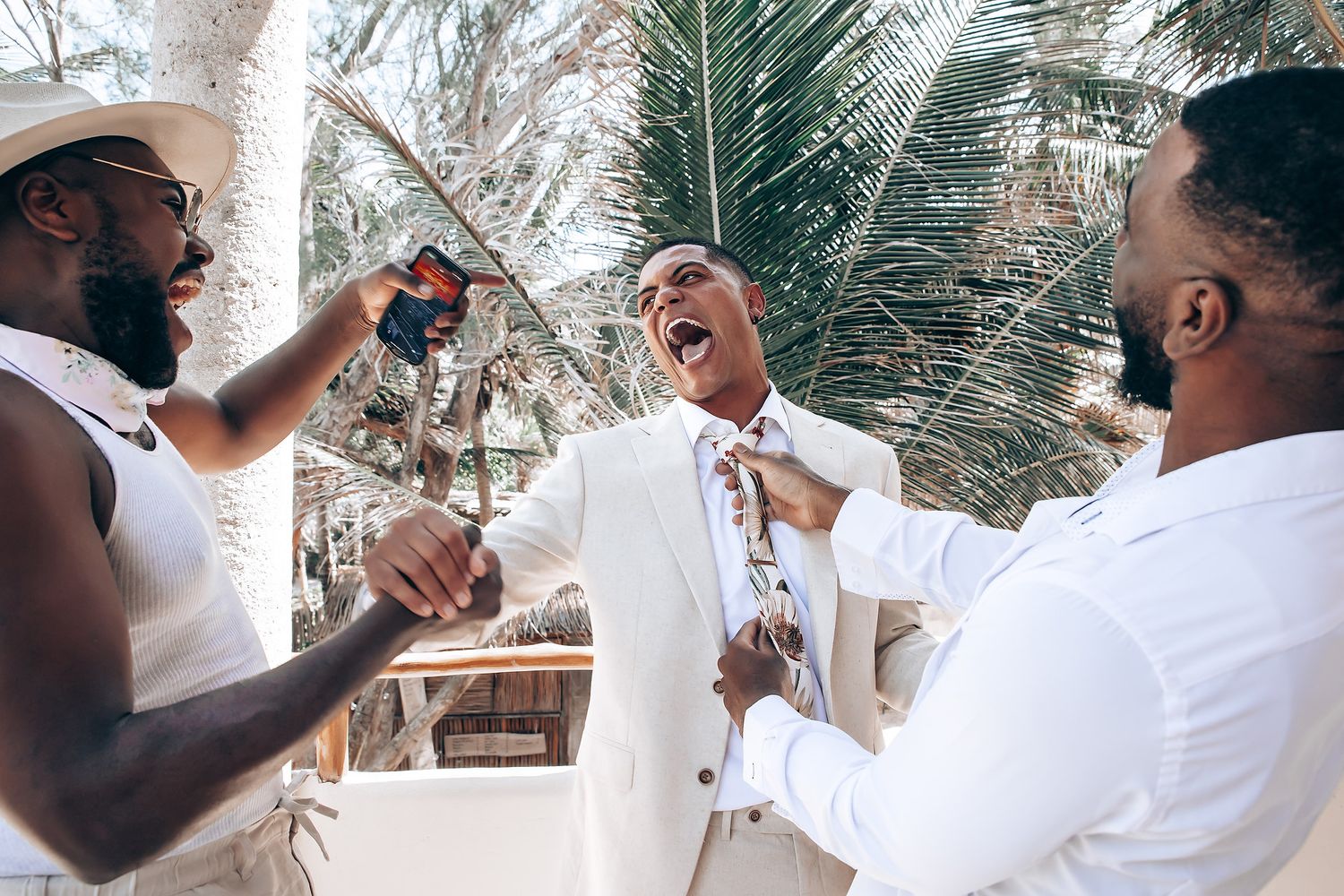 Joyful wedding party members share a candid laughing moment under palm trees.