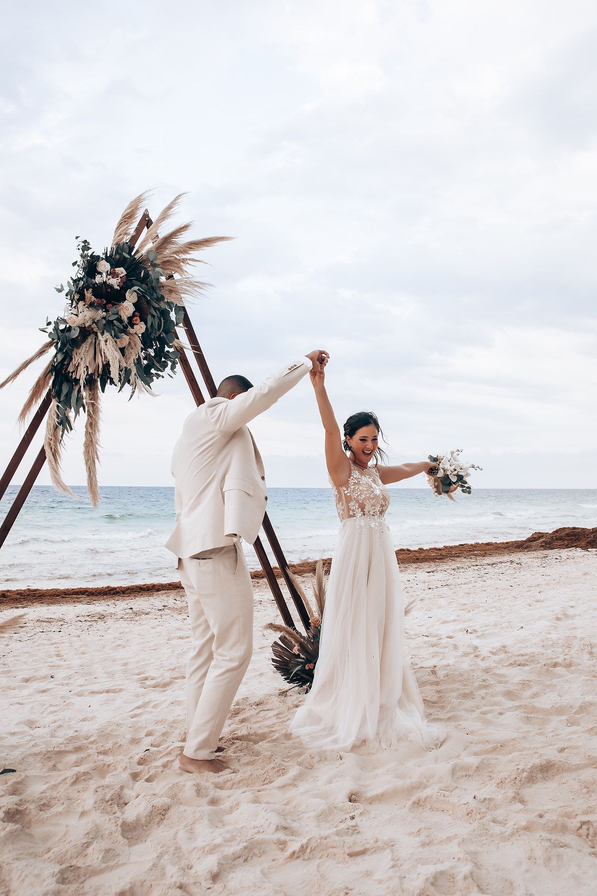 A celebratory moment under a bohemian beach wedding arch with dried flowers and pampas grass.