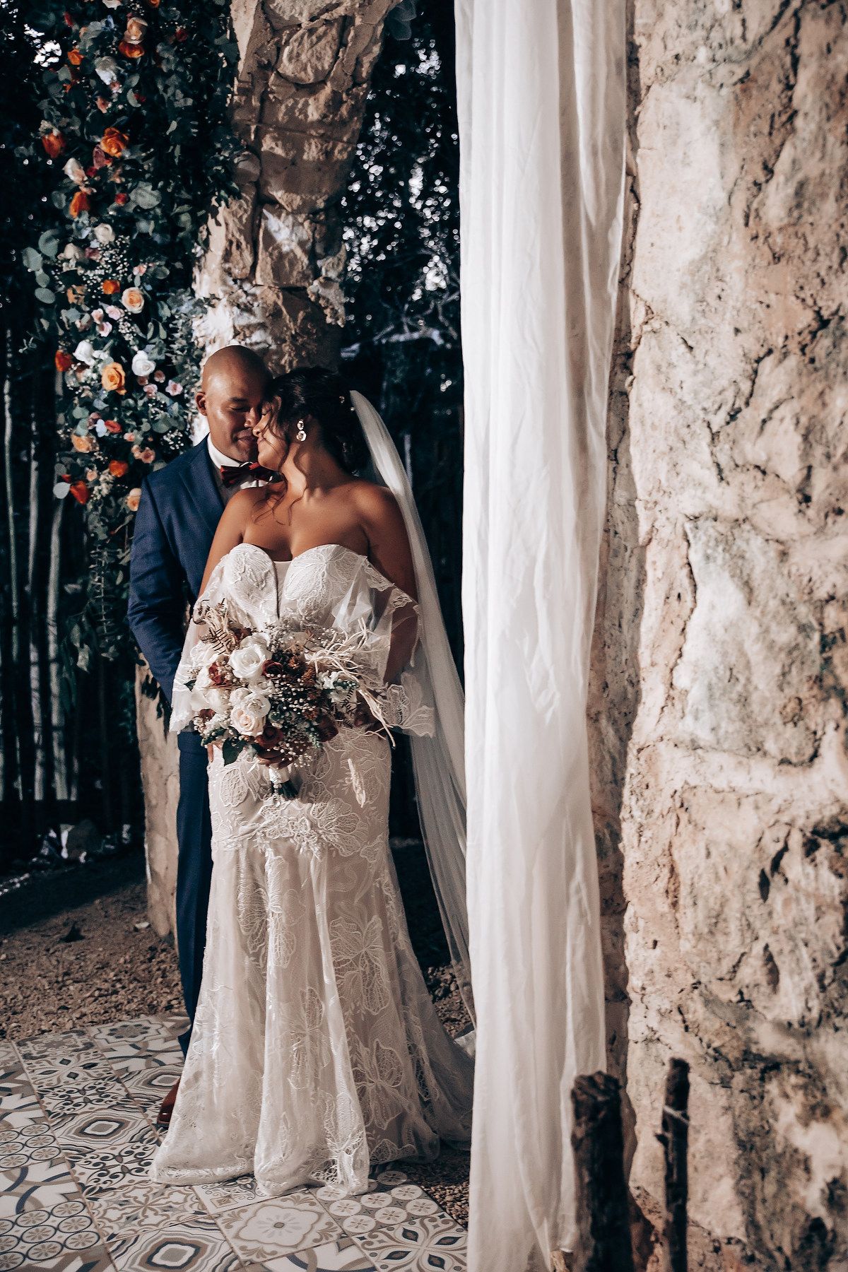 Elegant wedding portrait against stone wall with flowing white fabric and forest backdrop.