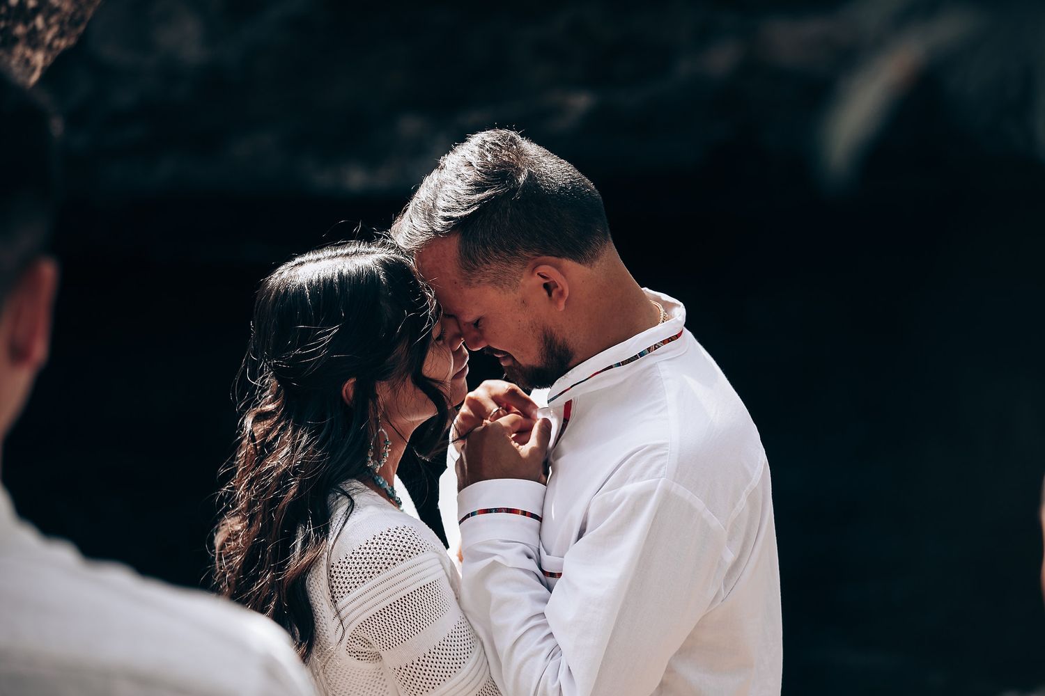 A tender embrace between a couple in white clothing against a dark moody background.