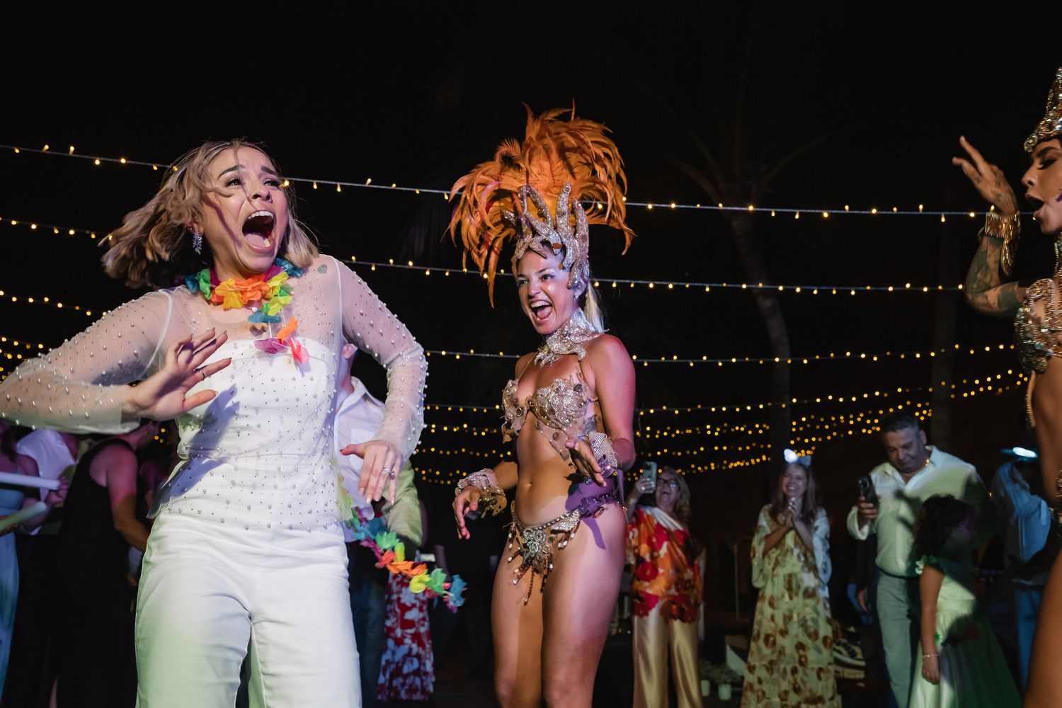 Two women dancing at a festive party, one in a white outfit with lei, another in a colorful carnival costume with feathered headdress.