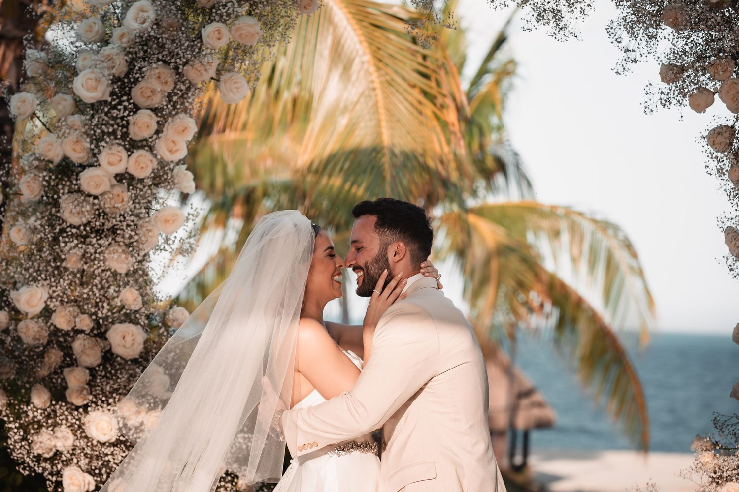 Bride and groom sharing first kiss under floral arch with palm trees and ocean backdrop.