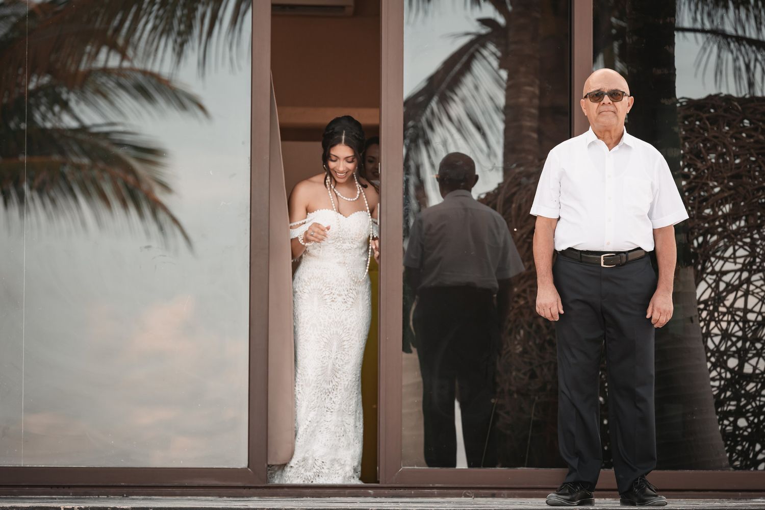 Bride in fitted lace gown exiting glass doors with older gentleman near tropical palm trees.