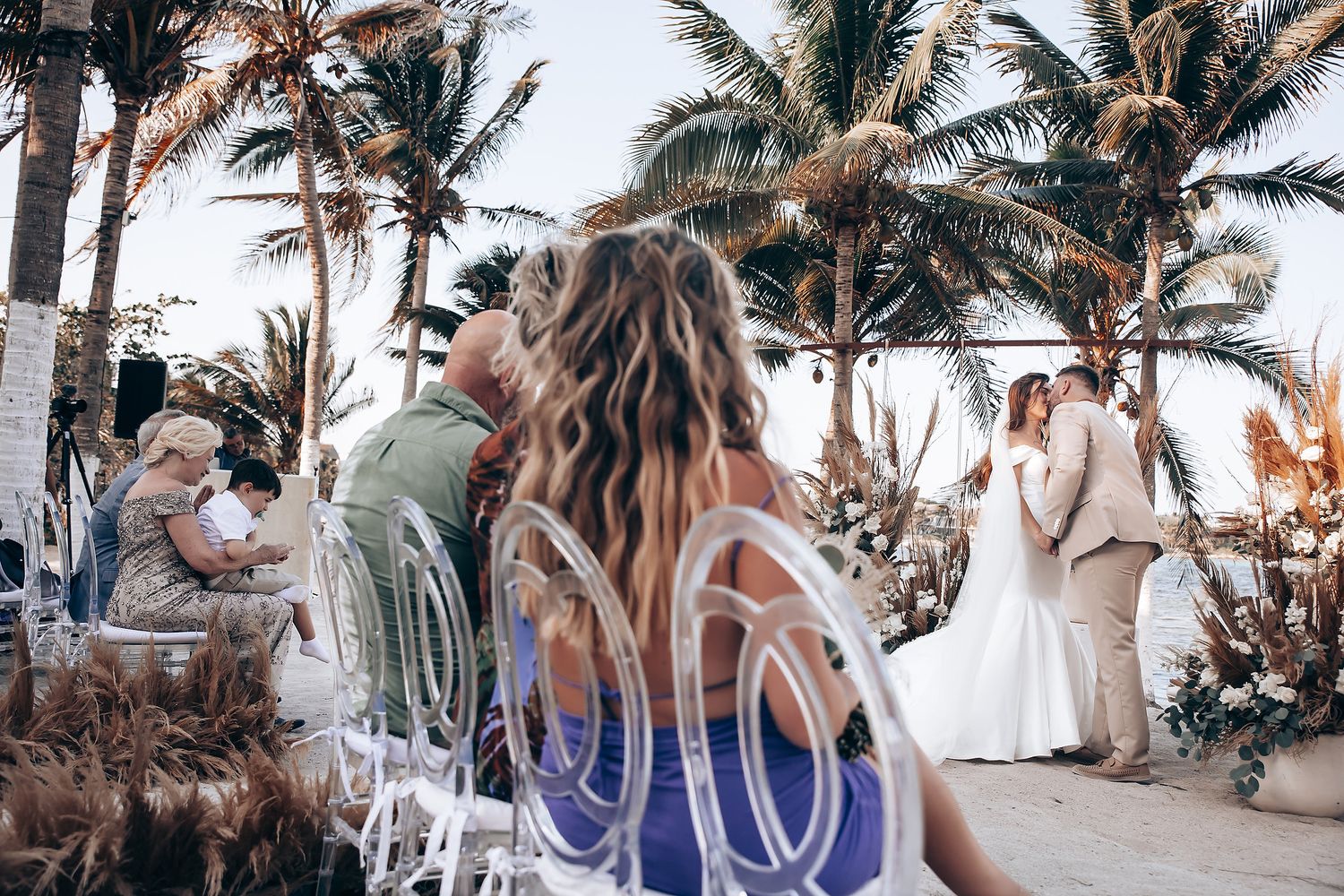 Beachfront wedding ceremony setup with white chairs and palm trees along the shoreline at dusk.