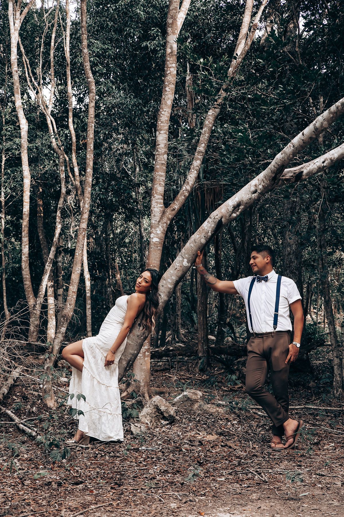 A couple in white and black outfits poses playfully against trees in a forest setting for romantic photos.