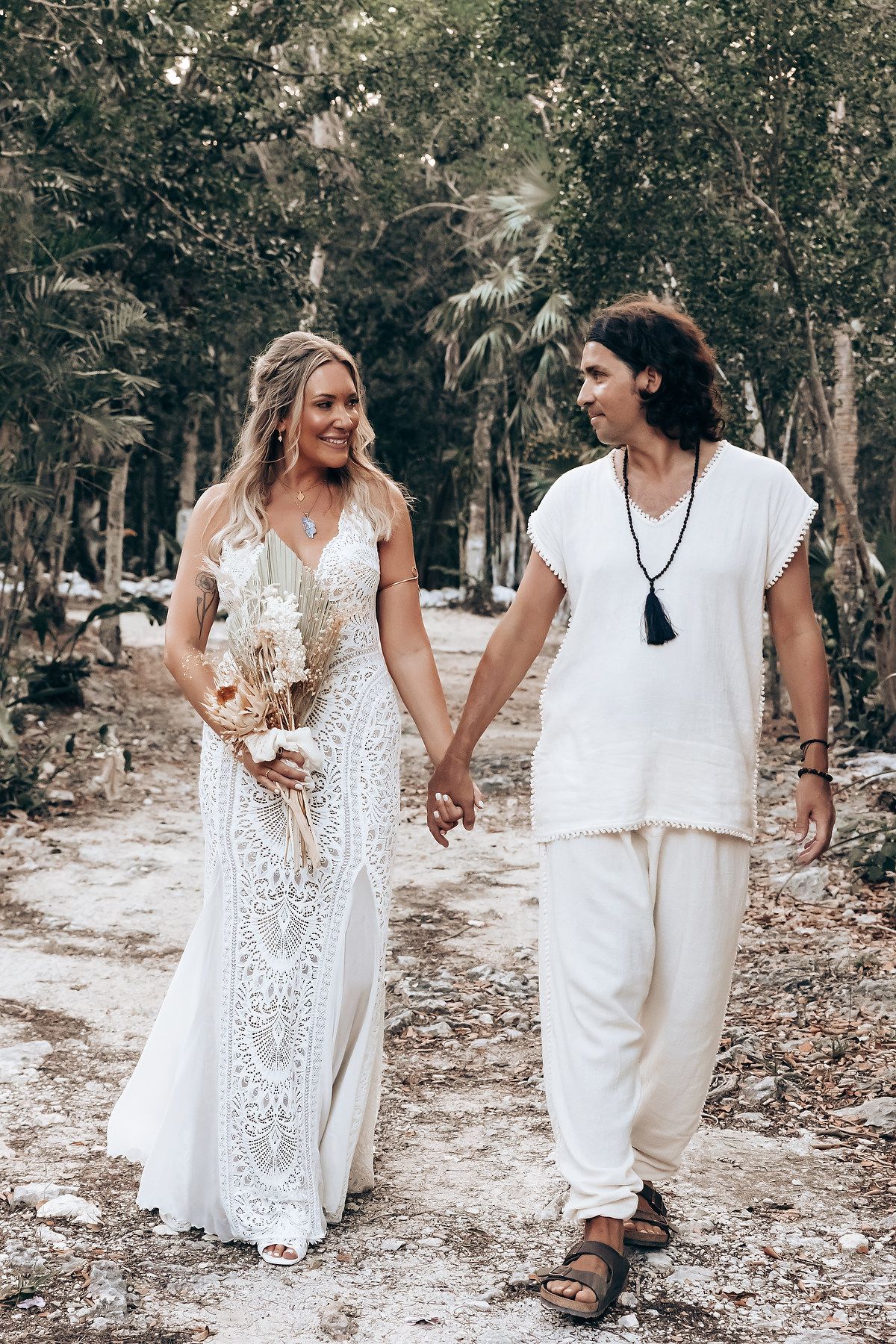 Two people in white outfits walk together and embrace on a wooded path during a romantic outdoor photoshoot.