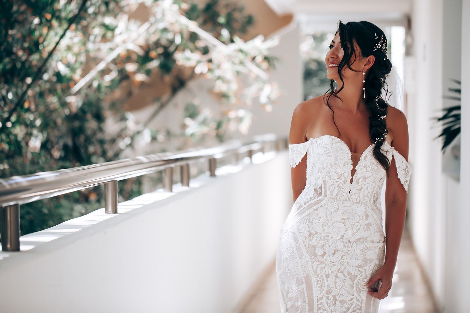 A bride in an elegant lace off-shoulder wedding dress stands on a white balcony surrounded by soft natural light.