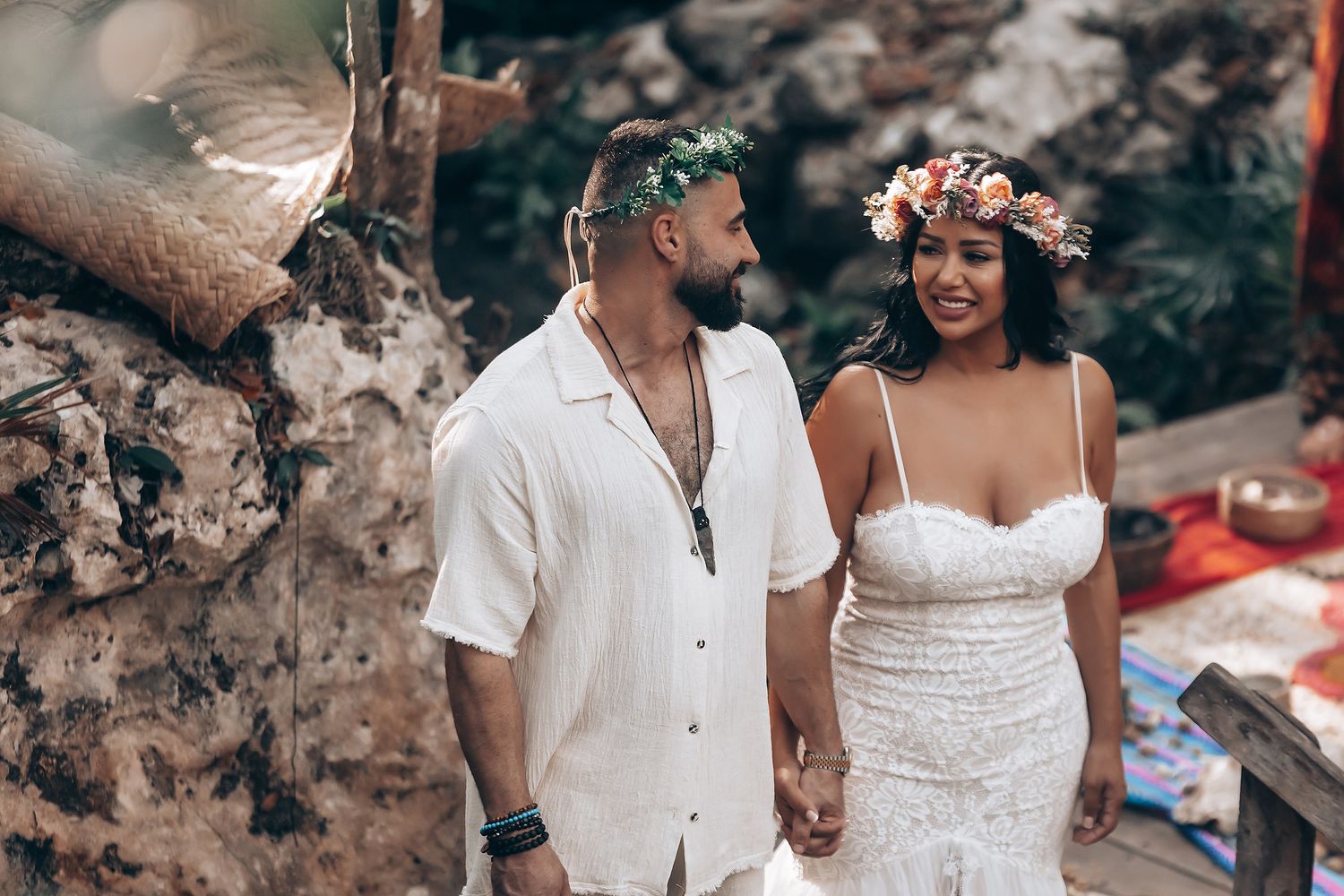 A couple in white wedding attire share an intimate moment in a rustic forest setting.