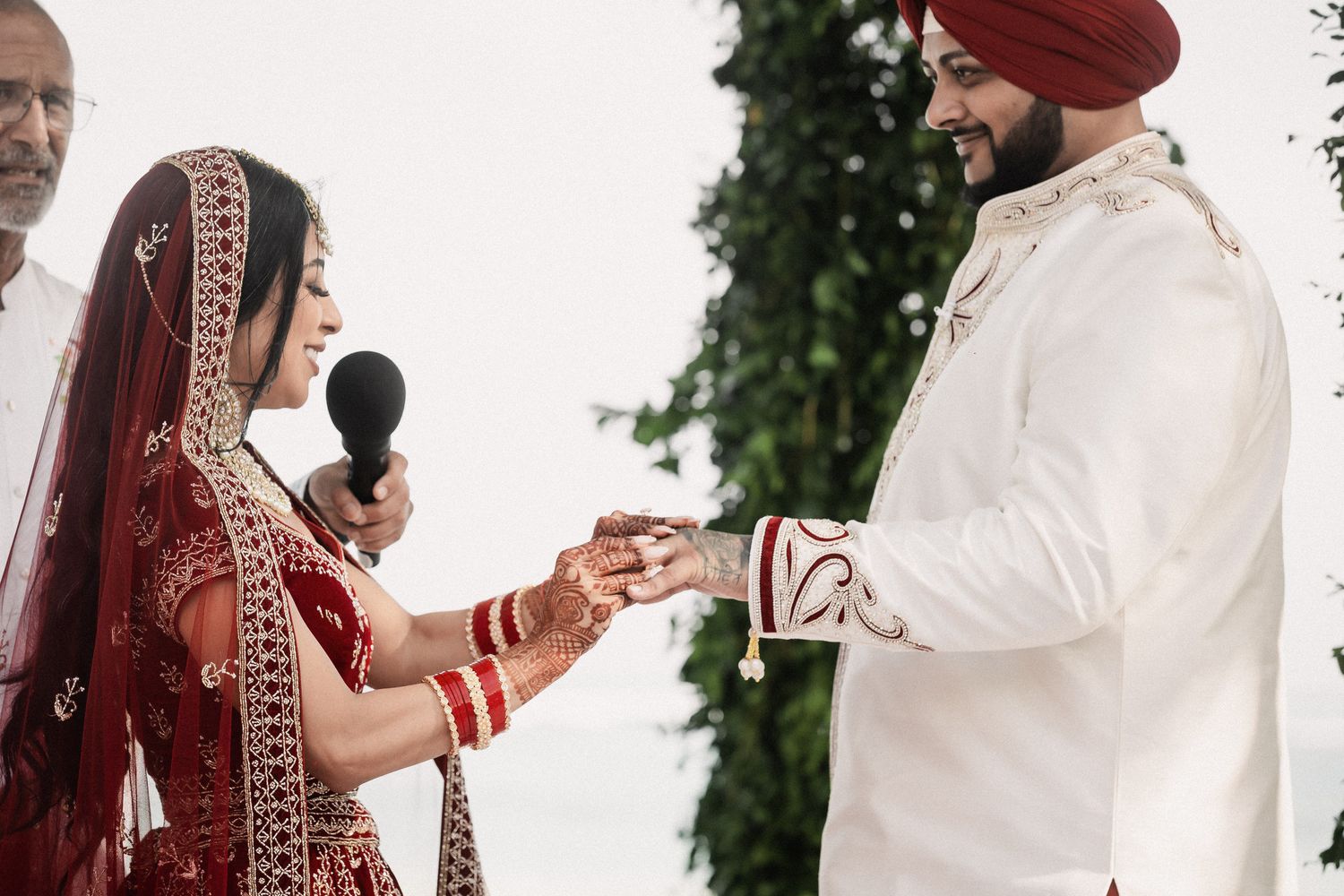 Indian bride in red lehenga placing ring on grooms finger during outdoor wedding ceremony.