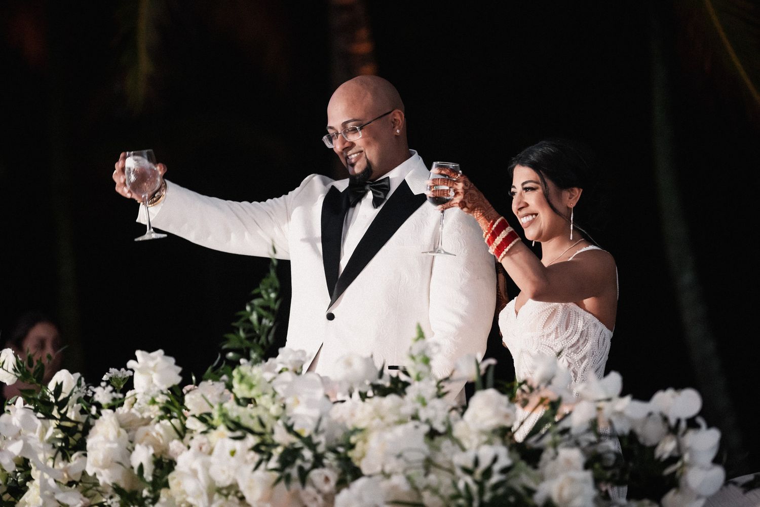 Elegant bride and groom toast with champagne glasses over white floral centerpieces at wedding reception dinner.