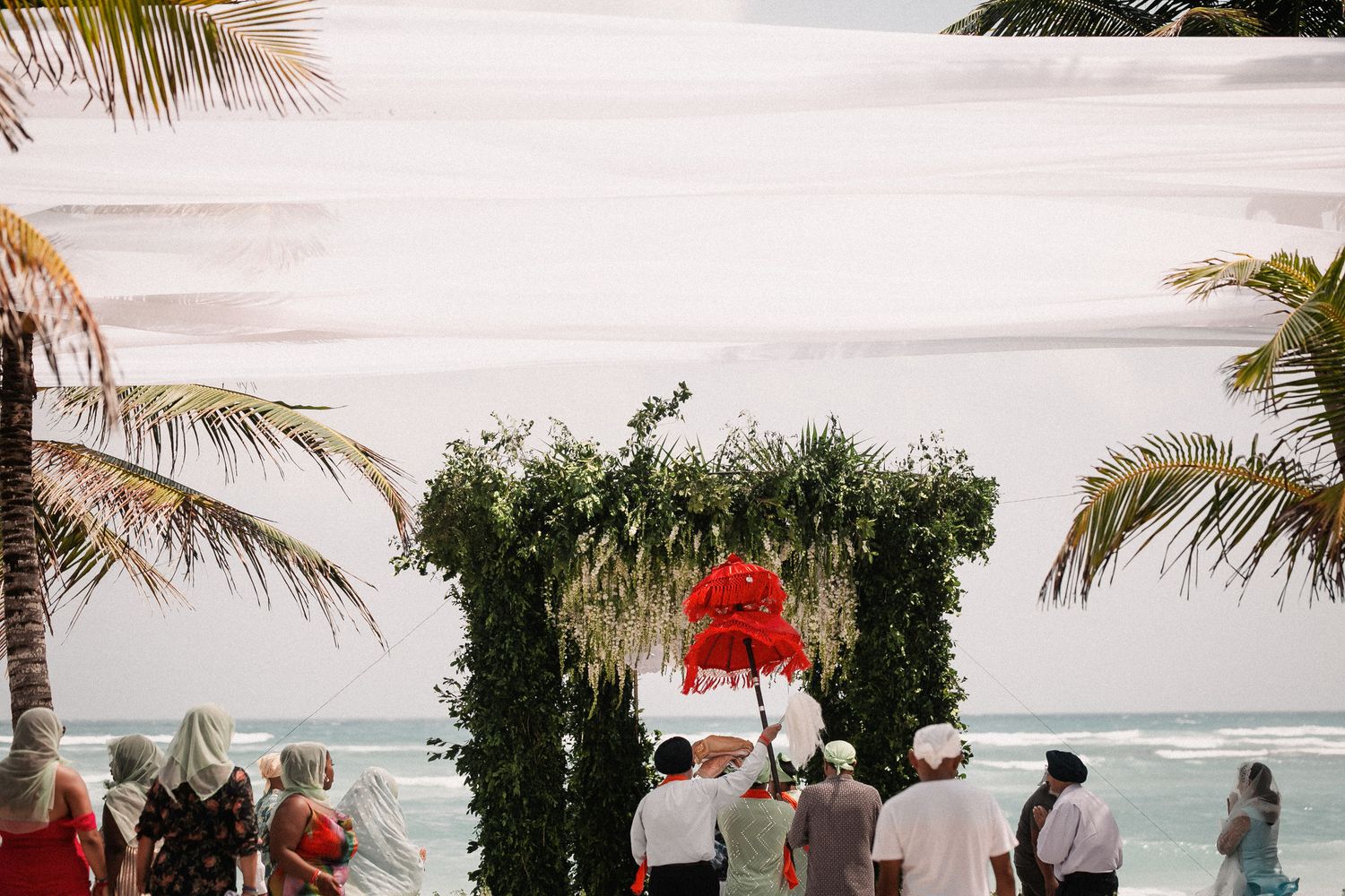 Outdoor beach wedding ceremony with floral arch decorated in greenery and red flowers by the ocean.