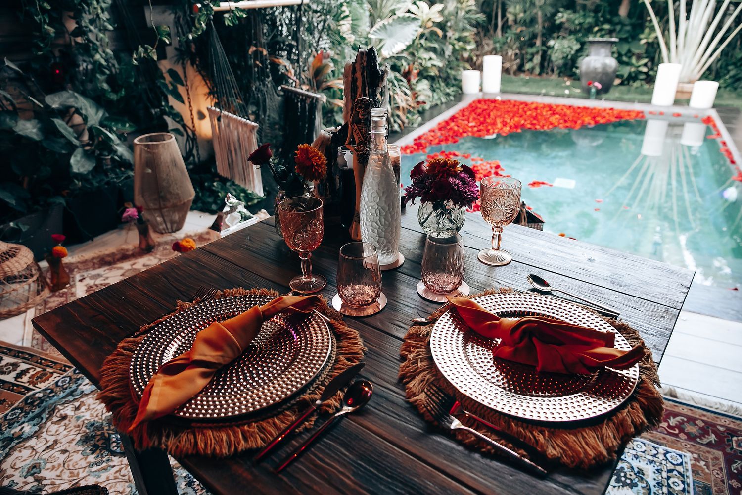 Elegant outdoor dining table set with woven placemats, burnt orange napkins, and glassware beside a flower-petal pool.