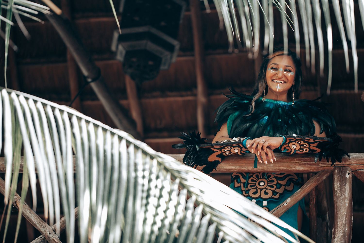 Smiling woman in teal feathered Mayan costume leaning on wooden railing surrounded by palm leaves.