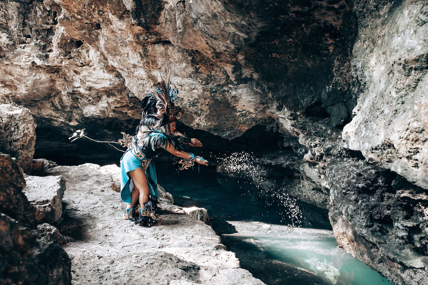 Person in Mayan ceremonial costume crouches over a turquoise cenote pool inside a rocky cave.