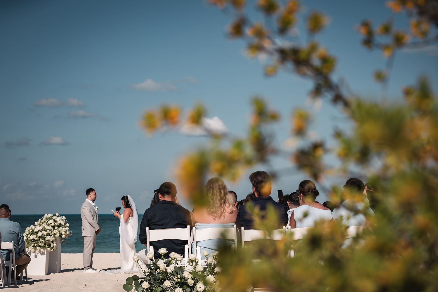 Guests seated at an elegant outdoor beach wedding ceremony with white floral arrangements and ocean views.