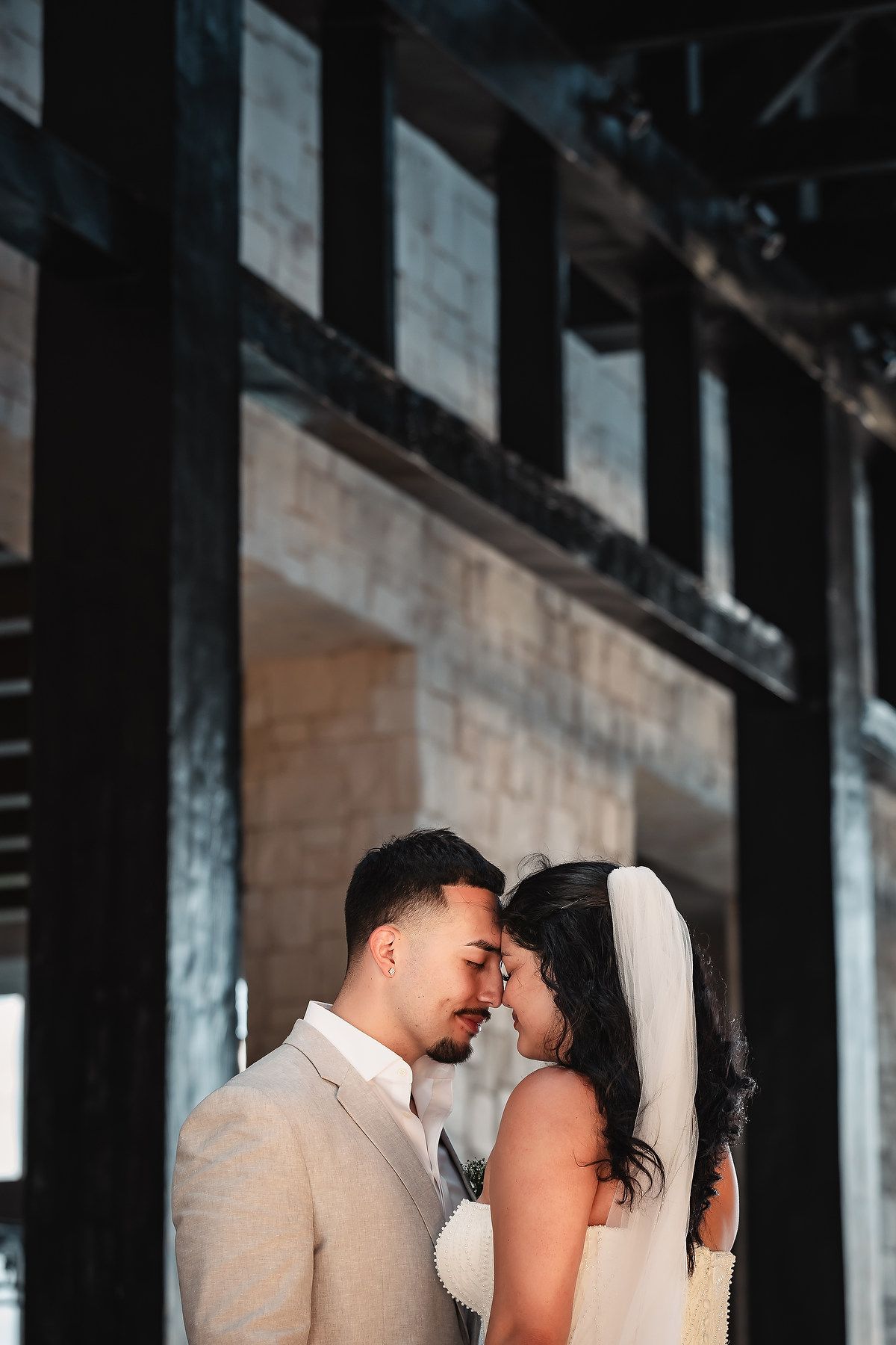 Bride and groom share an intimate moment under dark wooden beams, wearing white dress with veil and beige suit.