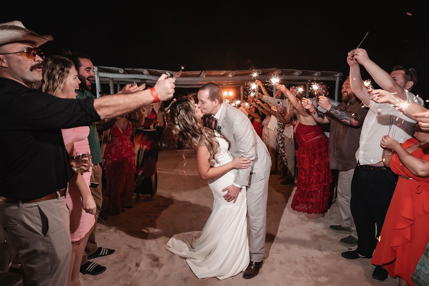 Bride and groom share a kiss during sparkler send-off surrounded by cheering wedding guests at night.