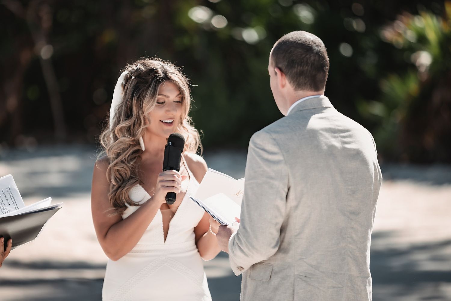 Bride smiling and reading wedding vows into a microphone while groom listens during outdoor ceremony.