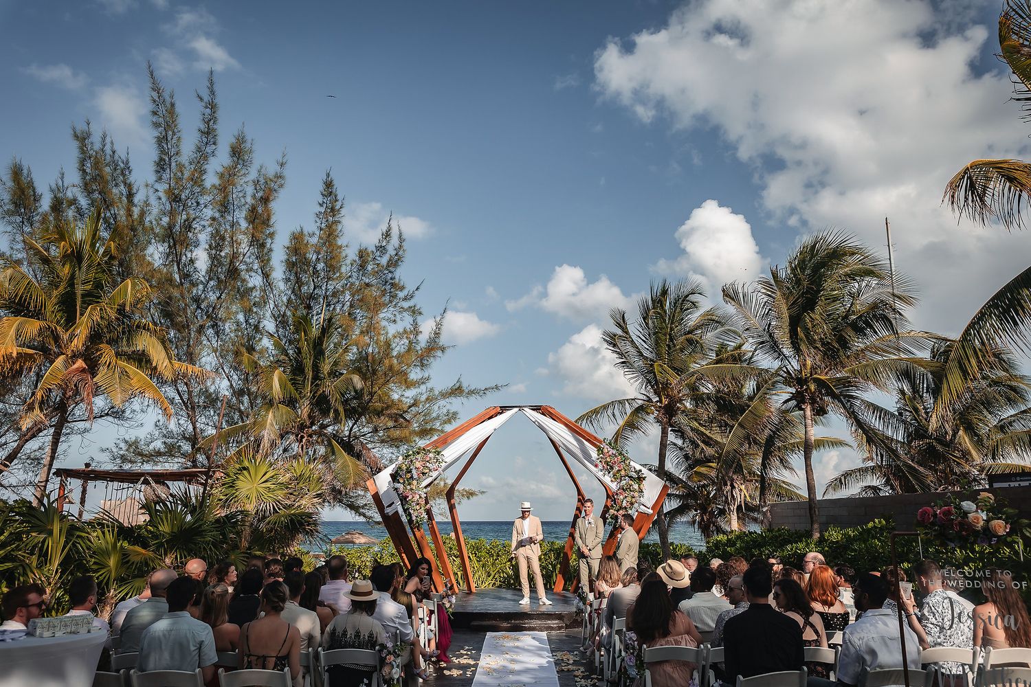 Outdoor beach wedding ceremony with guests seated under a colorful arch, surrounded by palm trees and blue skies.