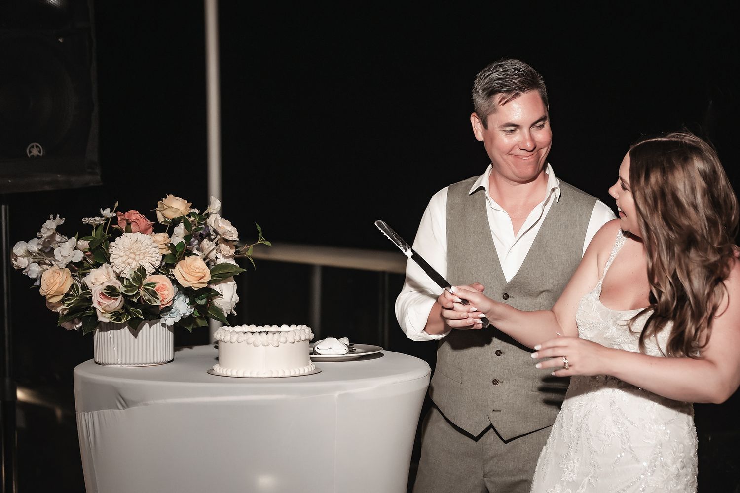 Bride and groom smiling while cutting wedding cake at elegant reception with floral arrangement.
