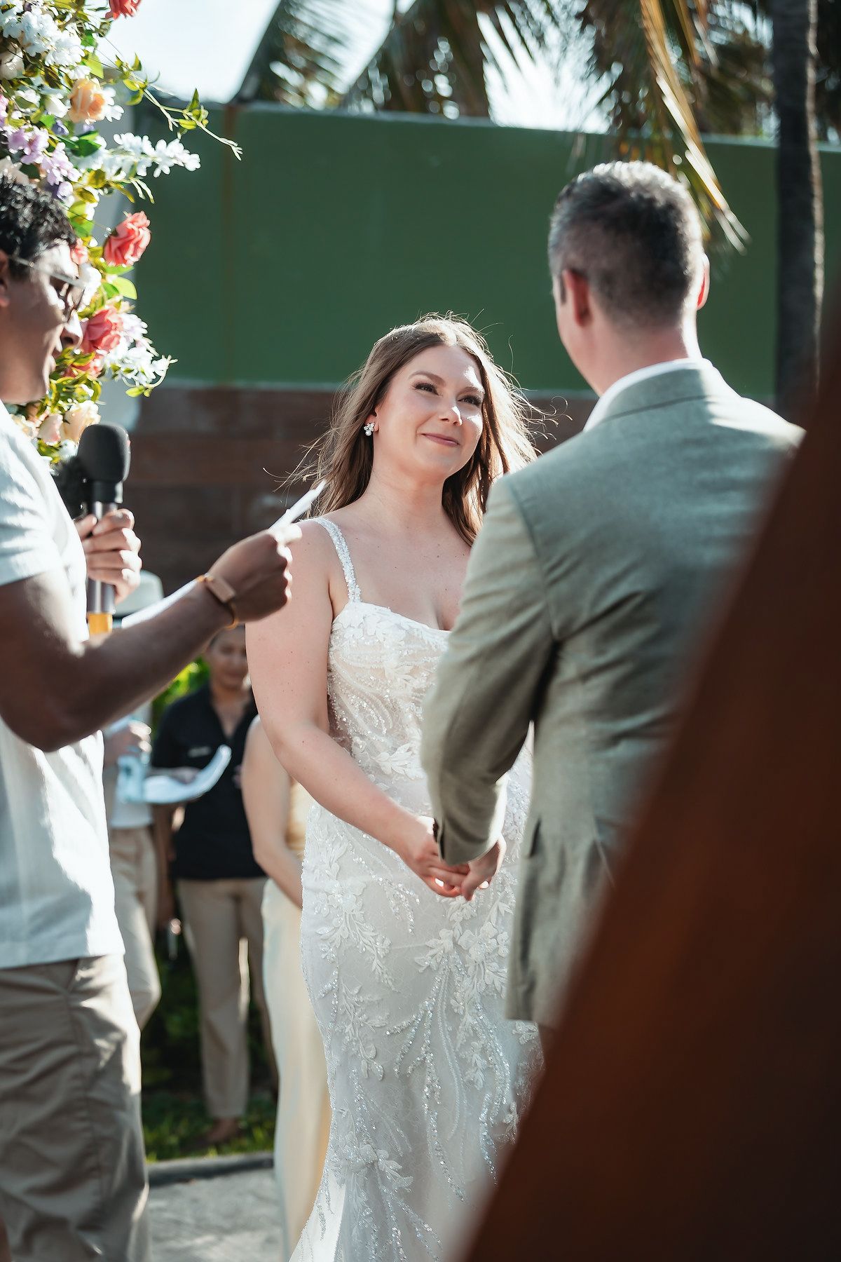 Bride in lace gown smiling at groom during outdoor wedding ceremony with officiant holding microphone.
