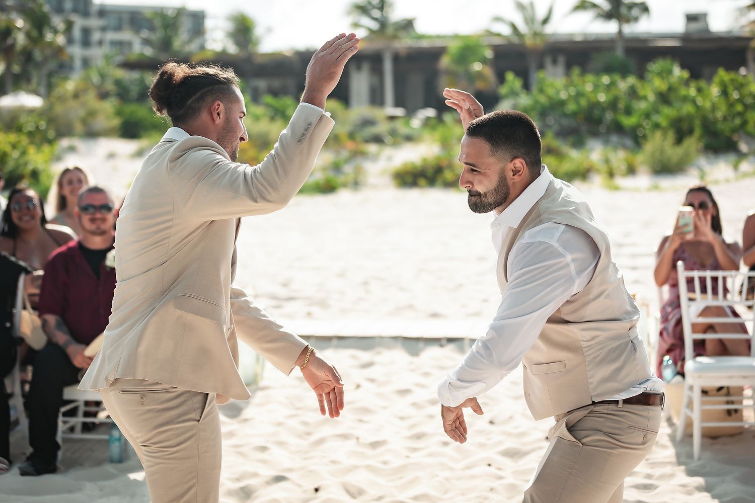 Two grooms in tan suits celebrate with a high-five dance at their beach wedding ceremony.