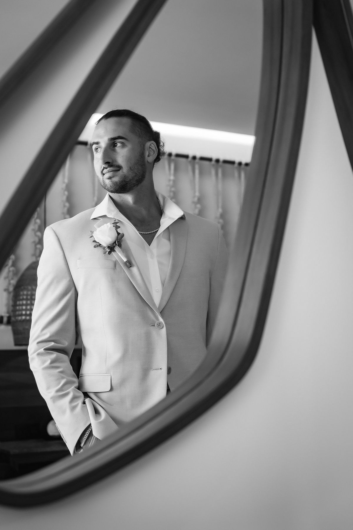Black and white photo of a groom in a light suit with boutonniere, reflected in a mirror, looking away thoughtfully.