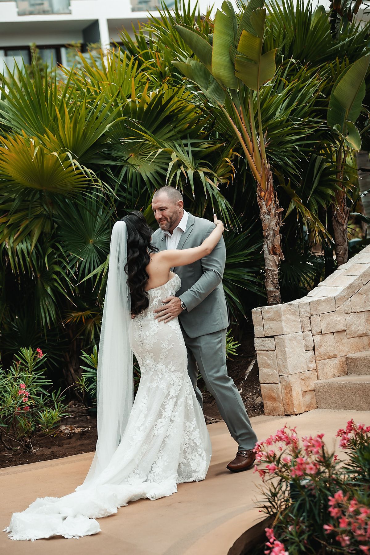 Bride and groom share a romantic embrace in a lush tropical garden, bride wearing a lace mermaid gown with veil.