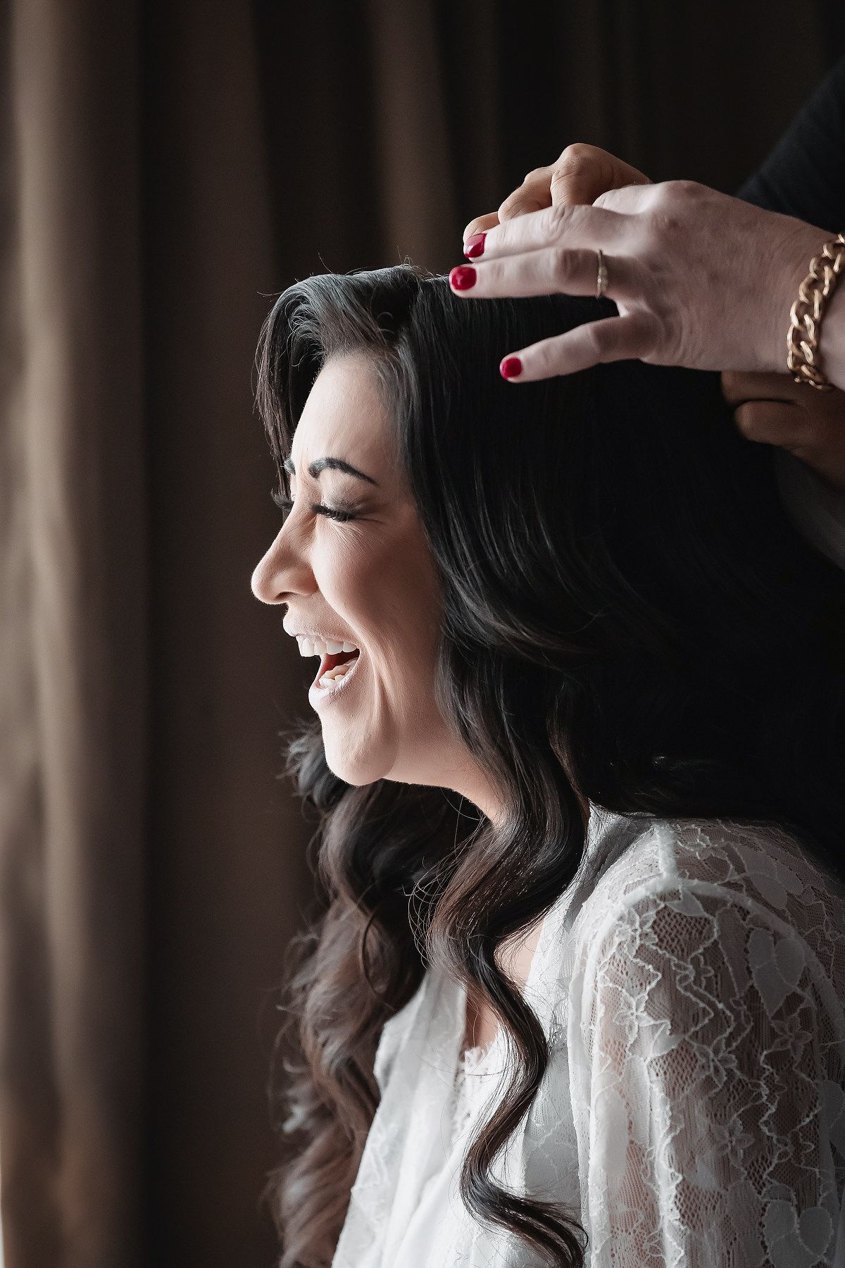 Bride laughing joyfully as hairstylist arranges her dark wavy hair, wearing a white lace robe during wedding preparations.