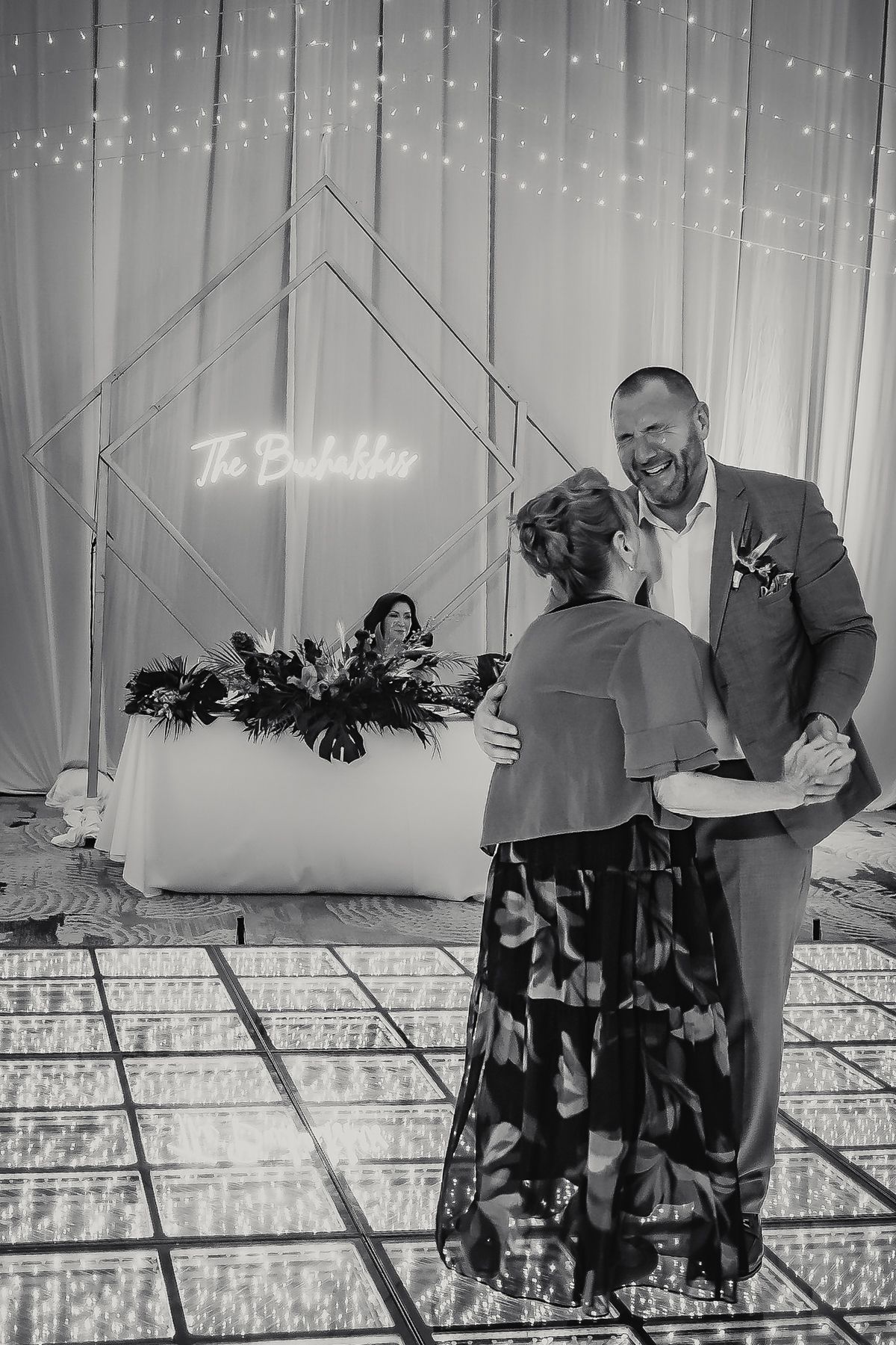 Black and white photo of a couple sharing a dance at a wedding reception with fairy lights and neon sign backdrop.