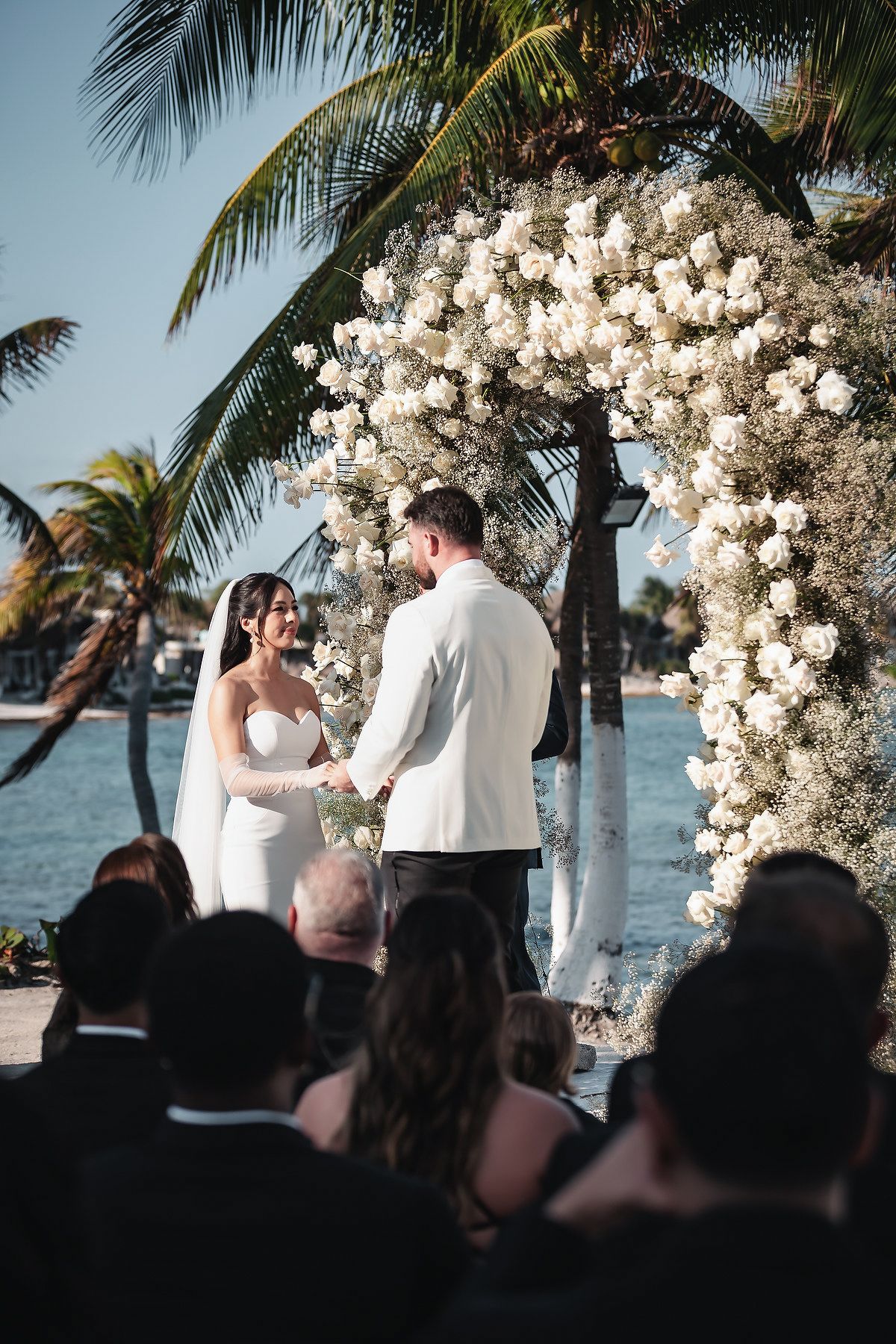 Bride and groom exchange vows under white floral arch by tropical waterfront with palm trees and seated guests.