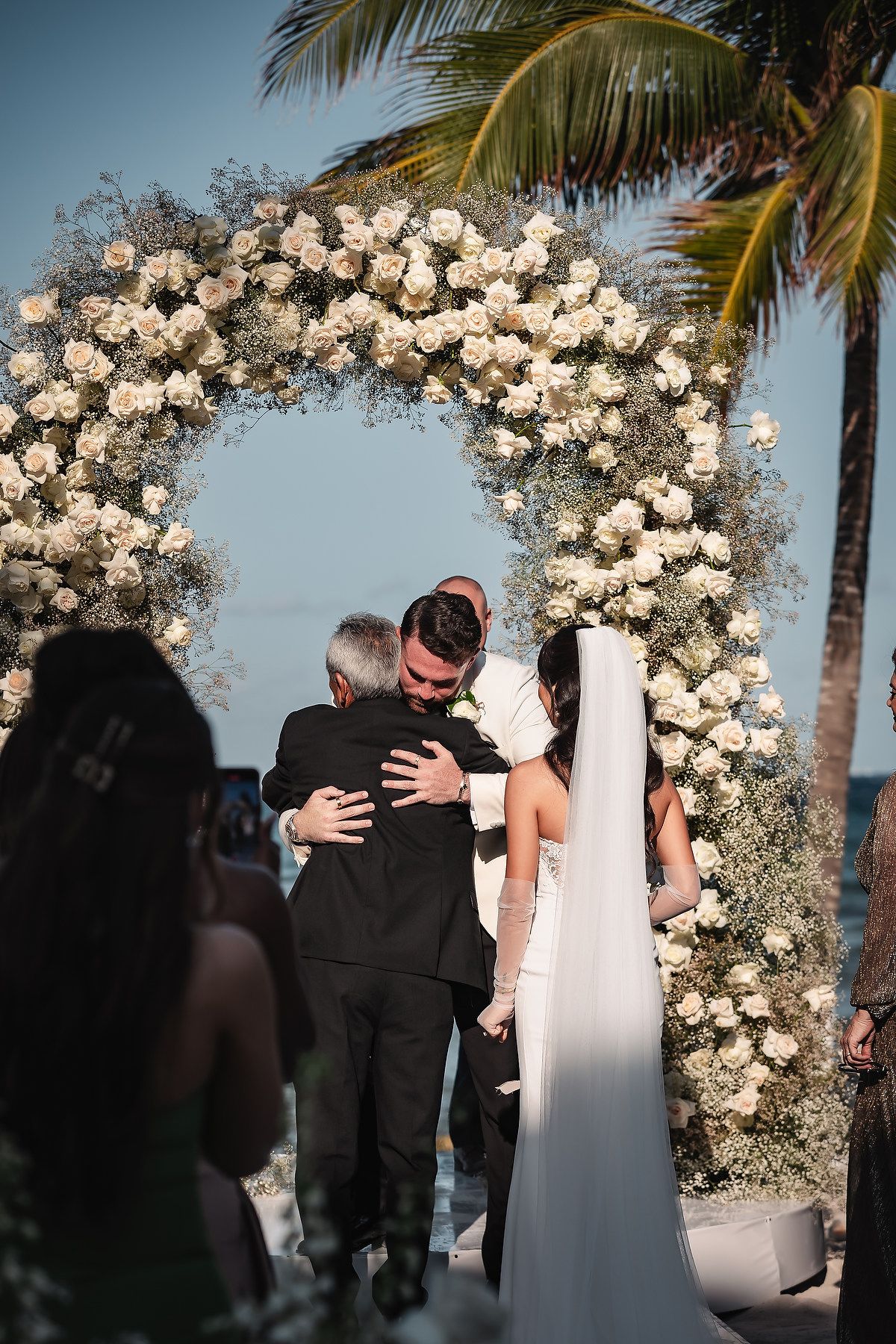 Groom hugs father-of-bride under stunning white rose floral arch at tropical beachside ceremony.