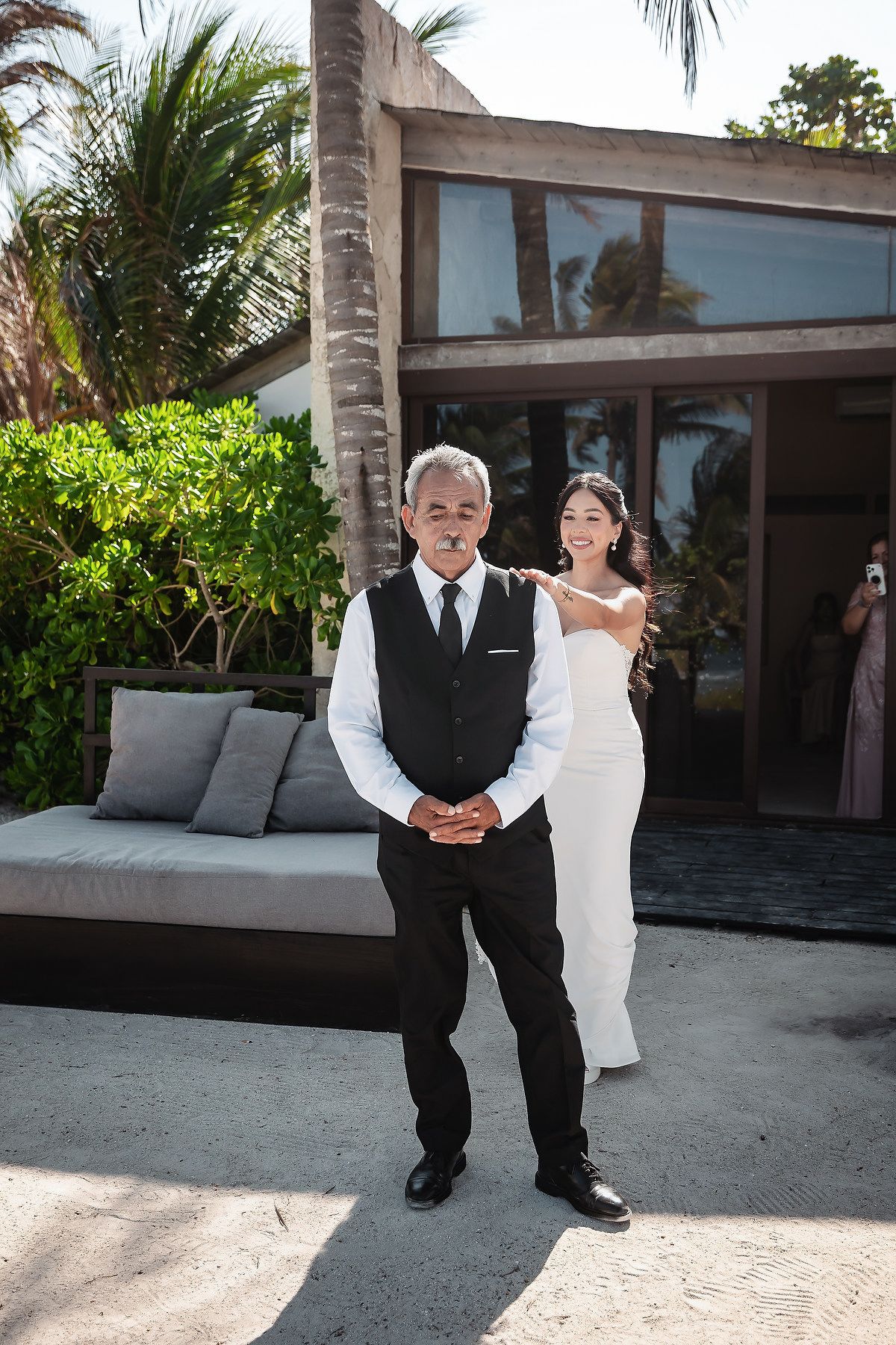 Bride in white gown stands behind older man in black vest and tie outside tropical villa with palm trees.