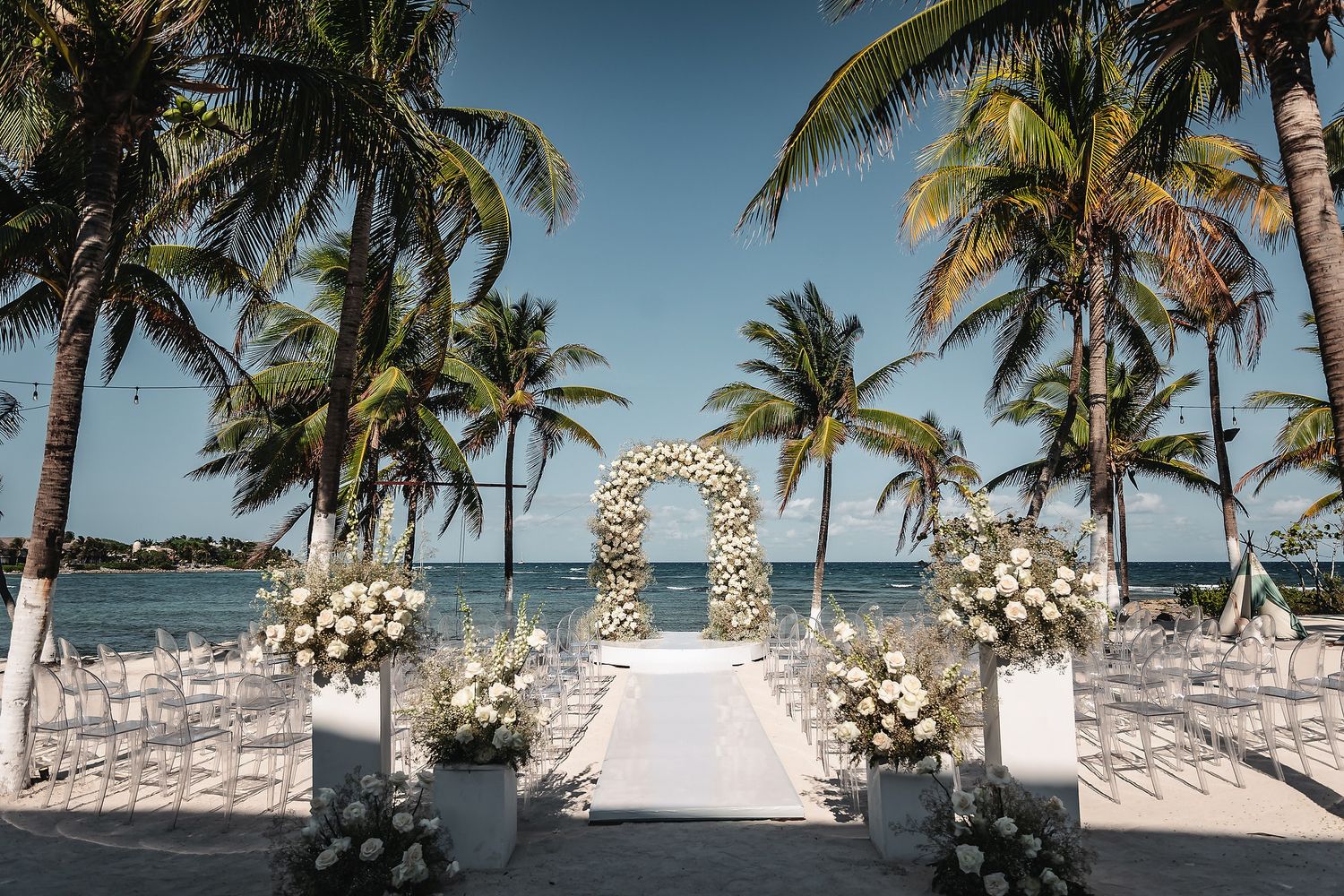 Elegant beachside wedding ceremony setup with floral arch, white chairs, and palm trees overlooking turquoise ocean waters.