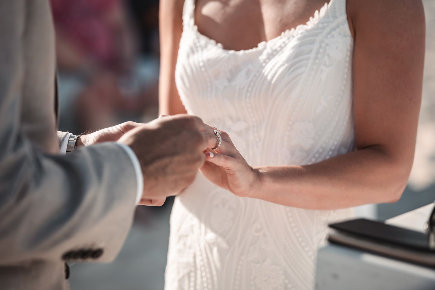 Bride in white lace dress receiving wedding ring from groom during outdoor ceremony.