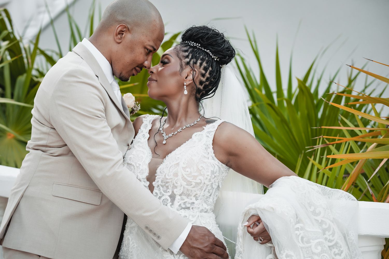 A bride and groom share an intimate moment, dressed in white lace gown and beige suit, surrounded by tropical greenery.