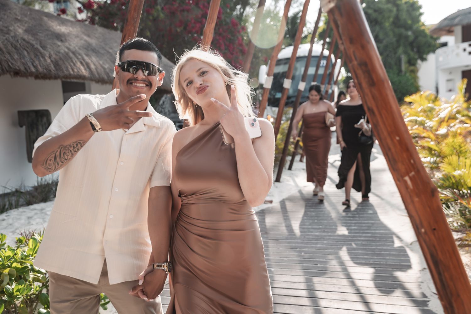 Couple walking together at a tropical resort, woman in brown dress and man in white shirt with sunglasses, guests in background.