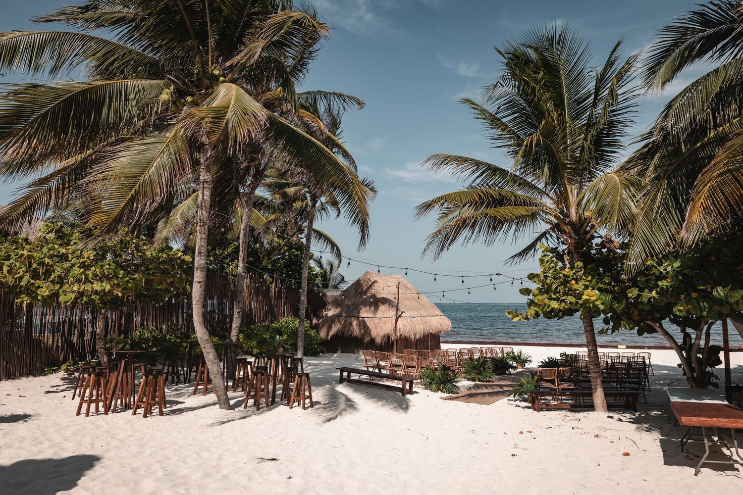 Tropical beach with white sand, palm trees, wooden chairs, and a thatched hut overlooking a calm blue ocean.