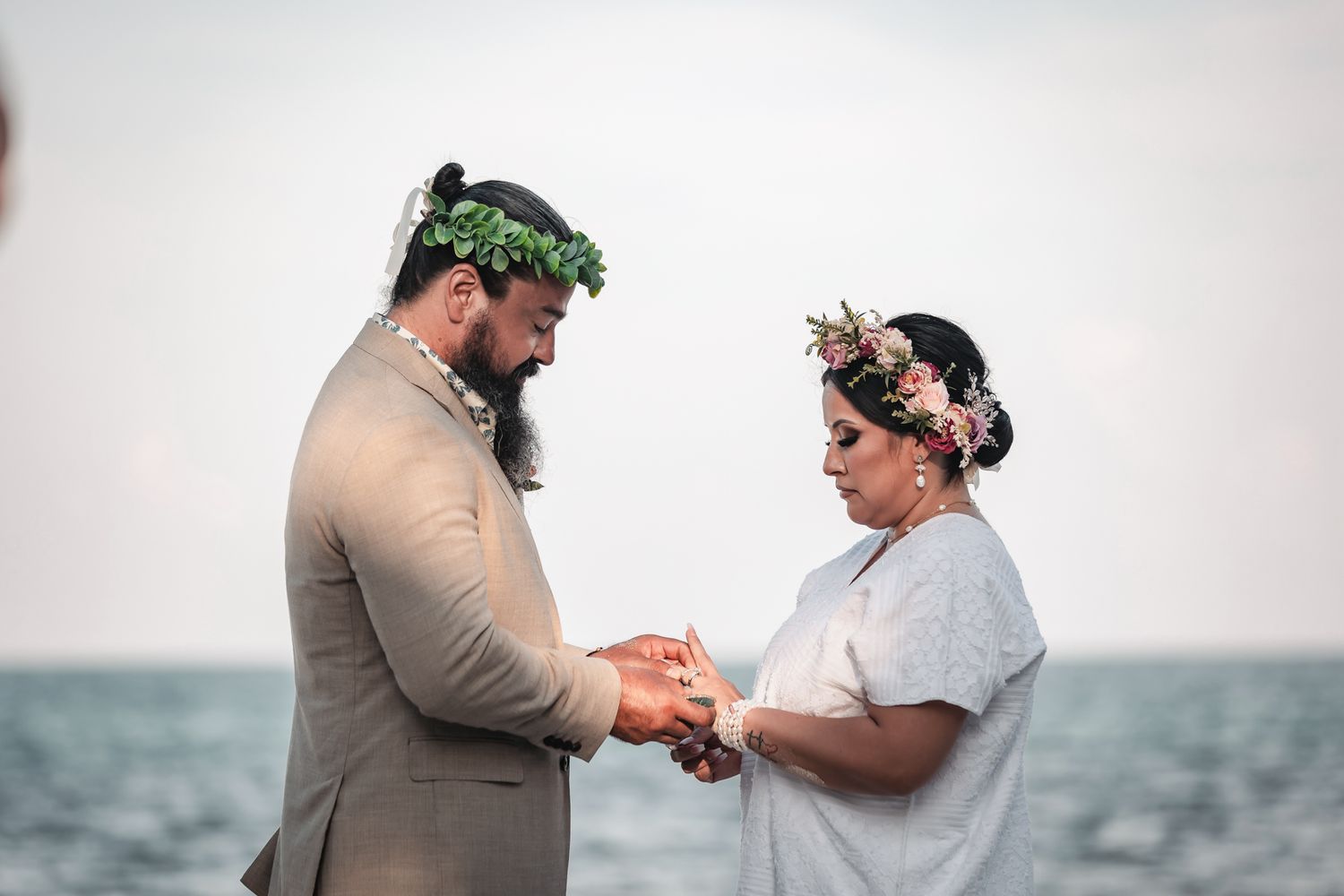 Couple exchanging rings at beach wedding ceremony, both wearing floral crowns, ocean backdrop.