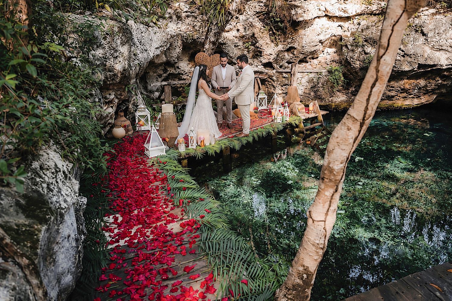 Romantic wedding ceremony in a lush cenote cave with red rose petal aisle and turquoise water backdrop.