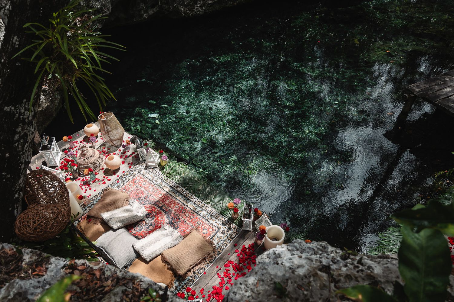 Romantic boho picnic setup on rocky cenote edge with red roses, woven rugs, and candles overlooking turquoise water.