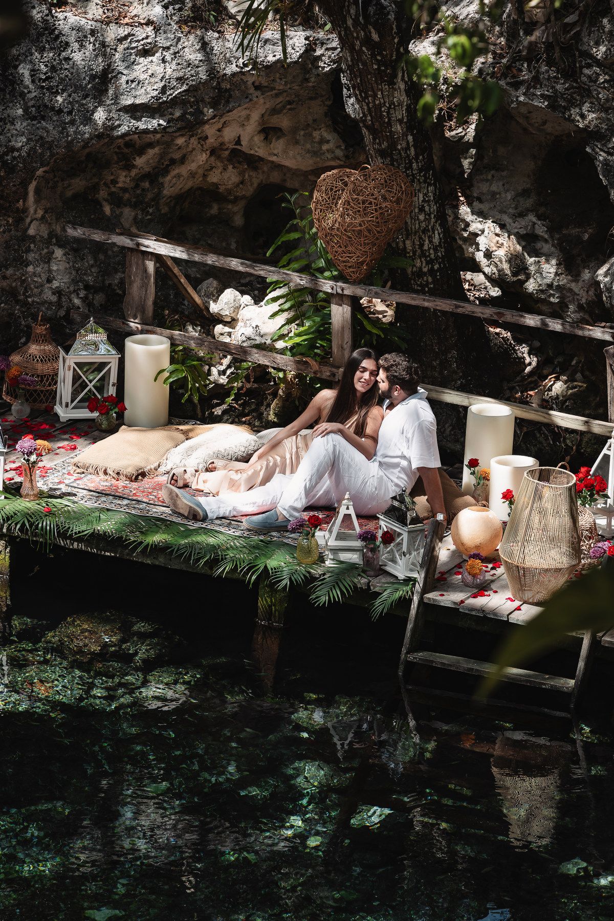 Romantic couple relaxing on a decorated platform over a cenote, surrounded by candles and tropical greenery.