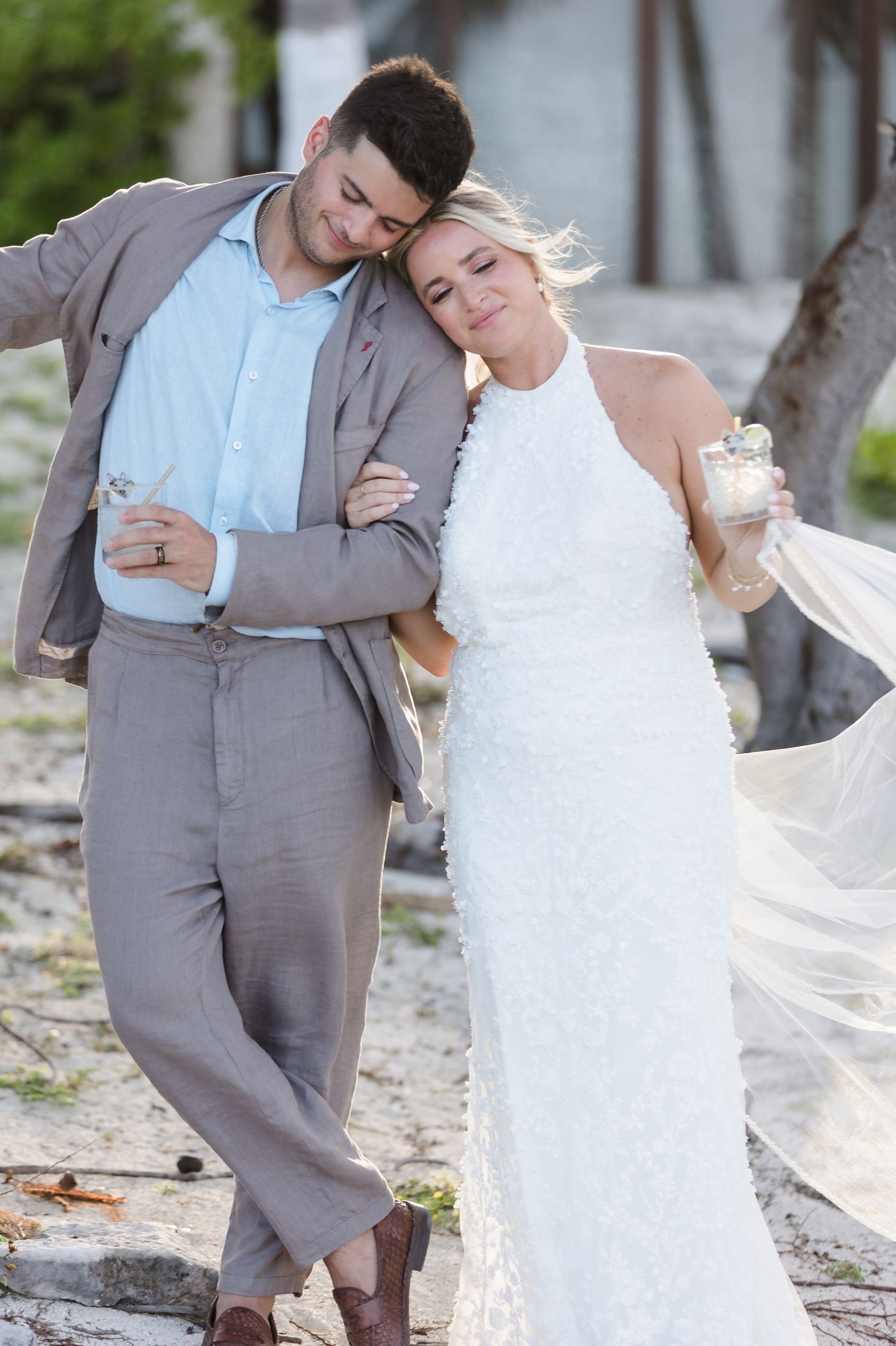 Bride in white halter gown leans on groom in gray suit during outdoor beach wedding celebration.
