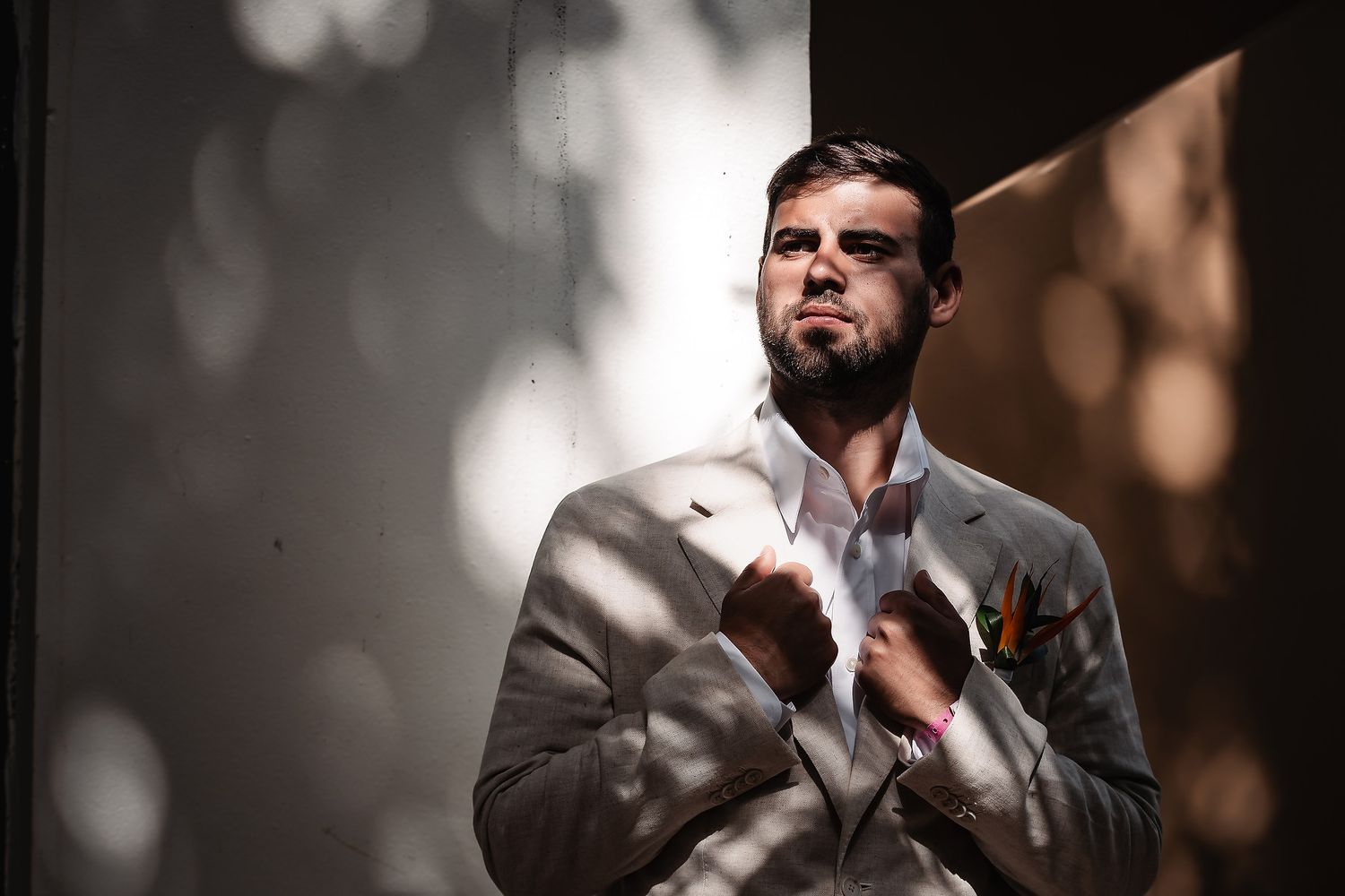 Elegant man in light gray suit adjusting tie in dramatic sunlight with bokeh background.