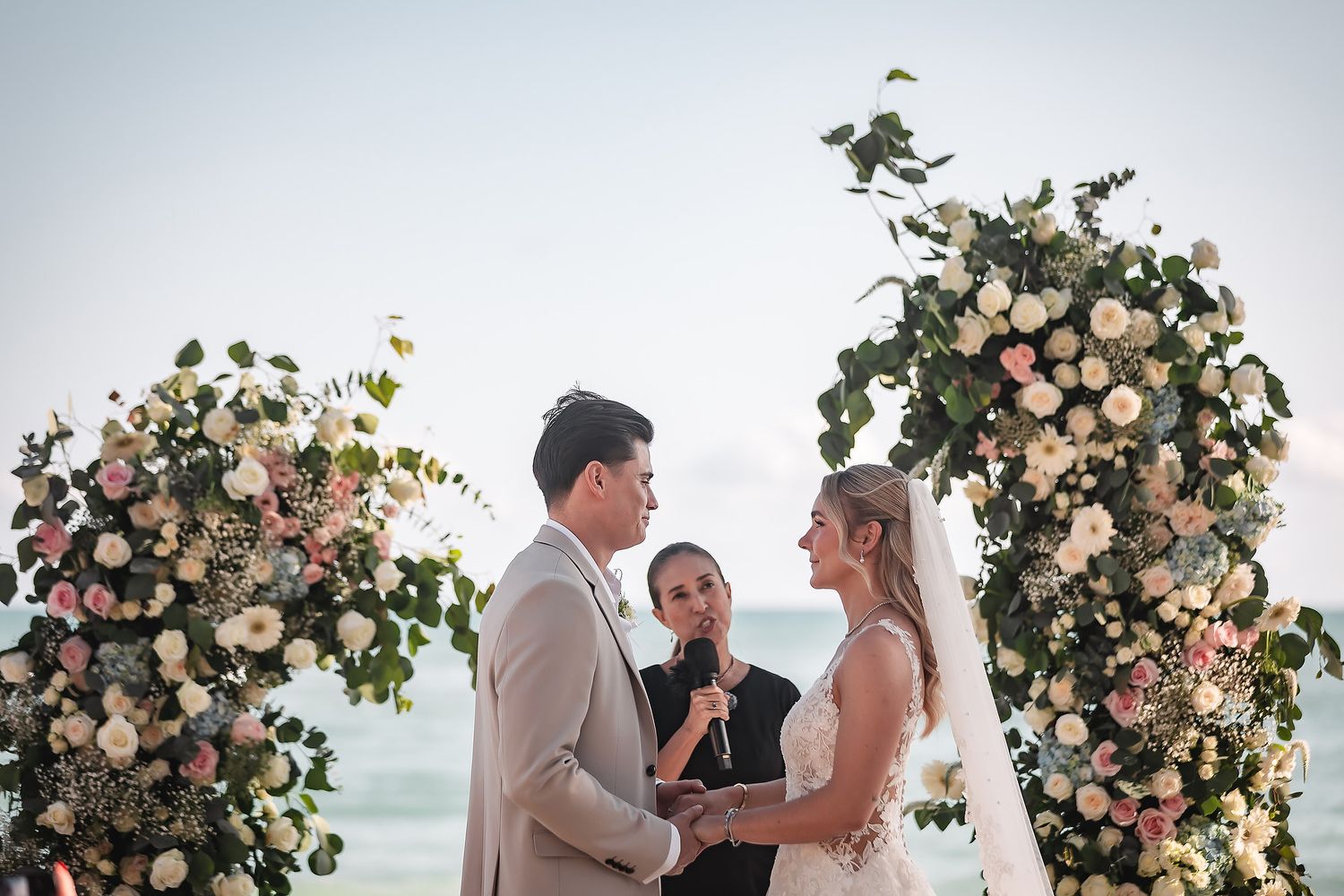 Intimate beach wedding ceremony under a floral arch with ocean views and palm trees.