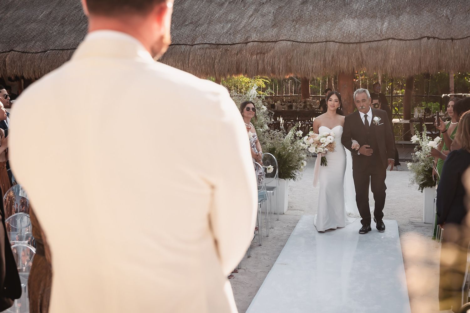 Wedding ceremony procession along a white aisle runner with tropical decor and palm trees in the background.
