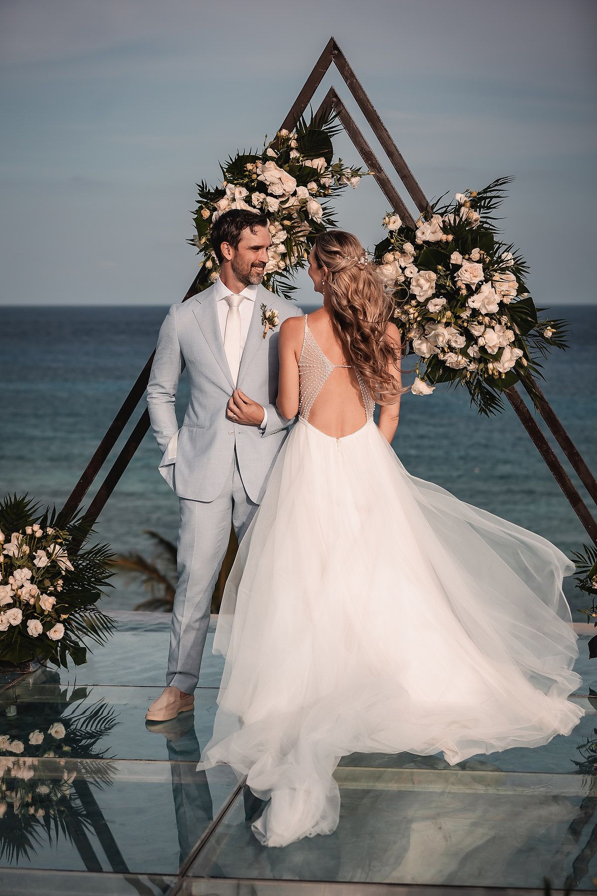 Seaside wedding ceremony featuring modern triangular arch decorated with blush roses.